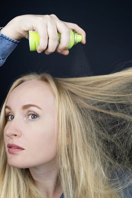 Une jeune femme blonde vaporise du shampoing sec sur ses cheveux